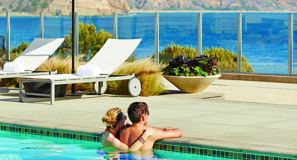 Two people are in a swimming pool, embracing, with lounge chairs, an umbrella, and a scenic ocean view in the background.