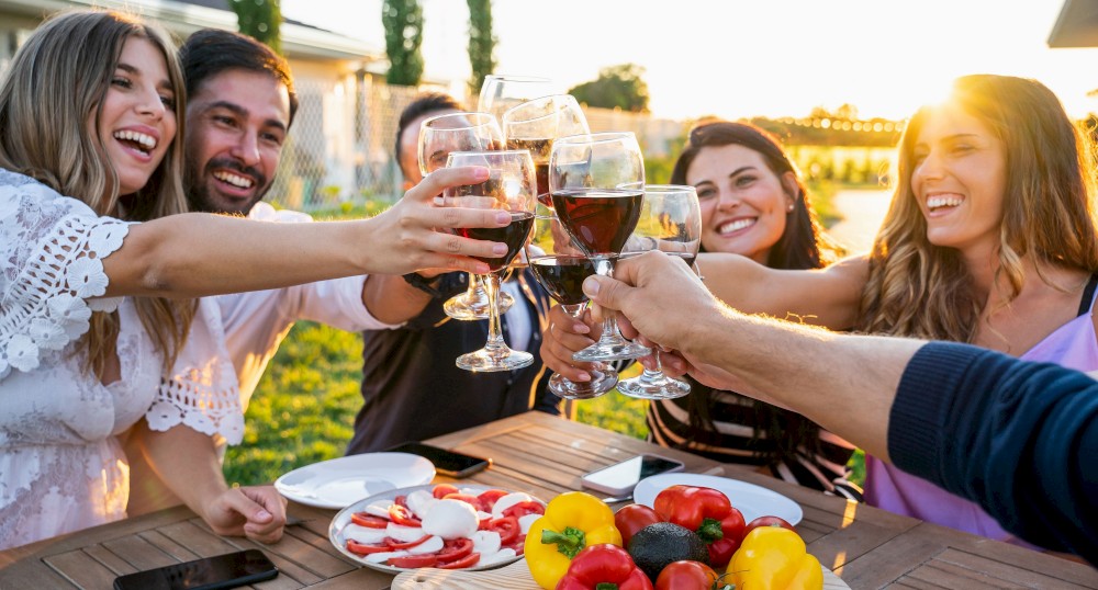 A group of friends clinks wine glasses around an outdoor table with food, featuring peppers, tomatoes, and cheese, during sunset.