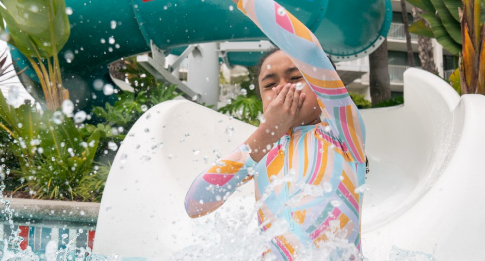 A child in a colorful swimsuit is sliding down a water slide, splashing into the pool below at an outdoor water park.