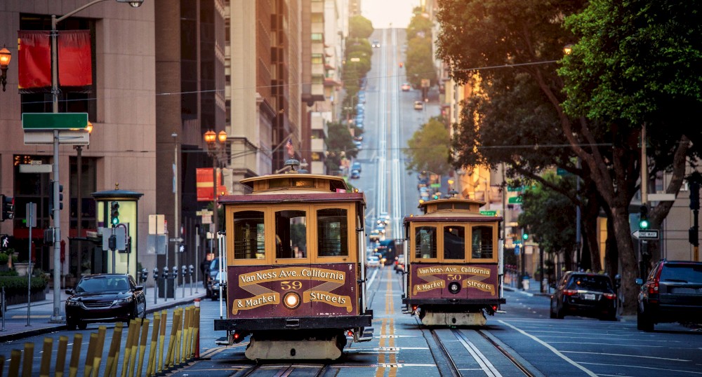 Two cable cars travel down a hilly urban street lined with tall buildings and trees, bathed in soft sunlight.