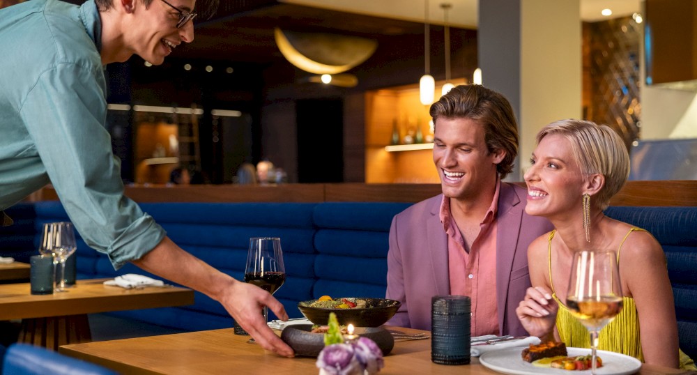 A waiter serves food to a smiling couple at a restaurant table, decorated with flowers and drinks, with a cozy and modern ambiance.