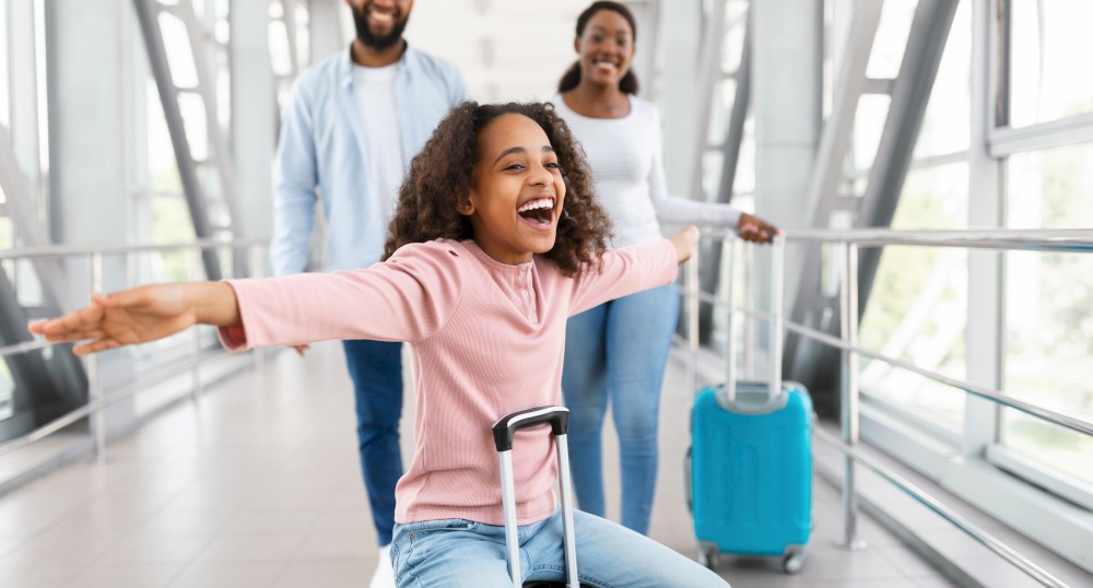 A joyful child rides on a suitcase at an airport, arms outstretched, while two adults follow behind with luggage, all smiling in a bright corridor.