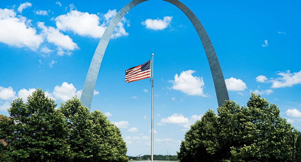 The image shows the Gateway Arch in St. Louis, Missouri with an American flag in the foreground, framed by green trees, under a clear blue sky.