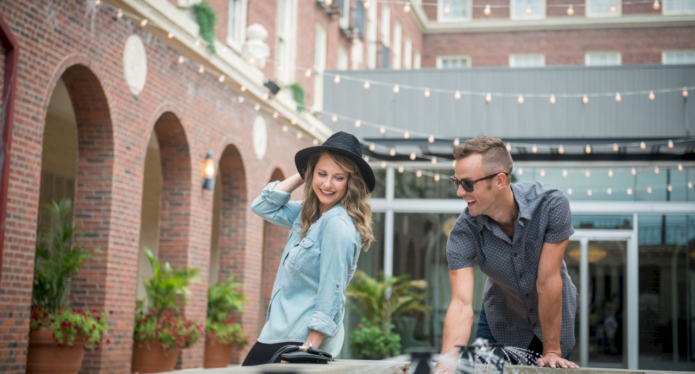 A woman and a man are enjoying themselves by a fountain in a courtyard with brick buildings and string lights, both smiling and casually dressed.