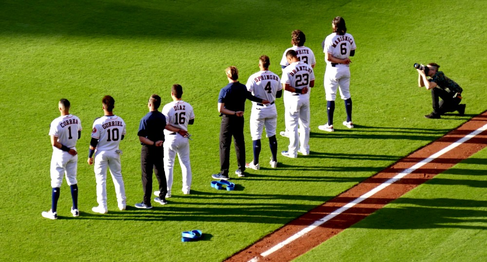 A group of baseball players is lined up on the field with a photographer capturing the moment, and some players have taken off their caps.