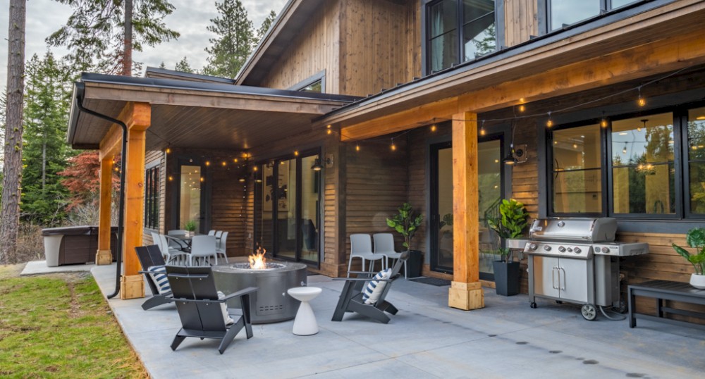 A cozy outdoor patio with seating, a fire pit, barbecue grill, string lights, and a modern wooden house in a forested area with a spacious lawn.