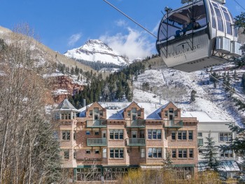 A gondola lift passes above a mountain resort with snow-covered peaks in the background and a large brick building in the foreground.