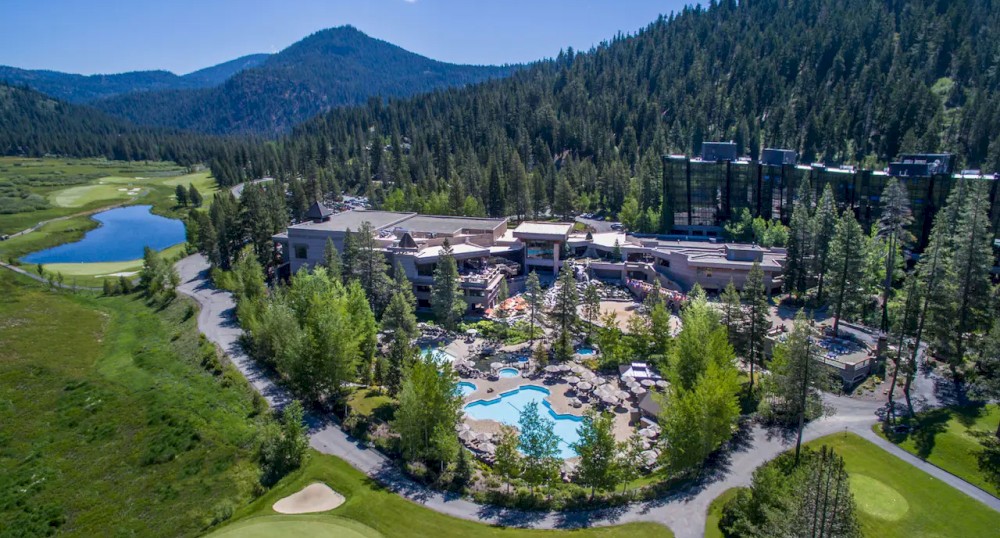An aerial view of a large resort complex nestled in a forested area, featuring several buildings, a swimming pool, and a golf course, surrounded by mountains.