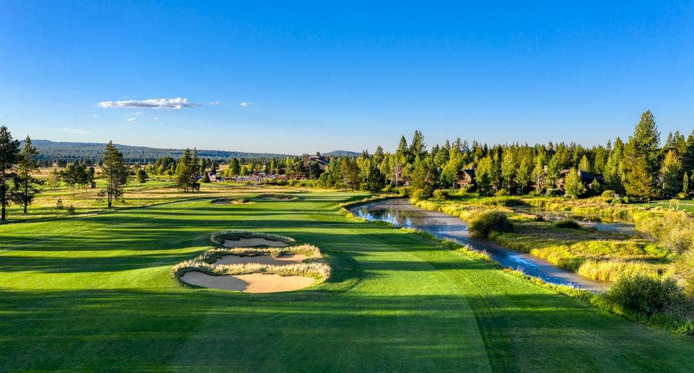 The image shows a scenic golf course with sand bunkers and a stream running through it, surrounded by lush greenery and trees under a clear blue sky.