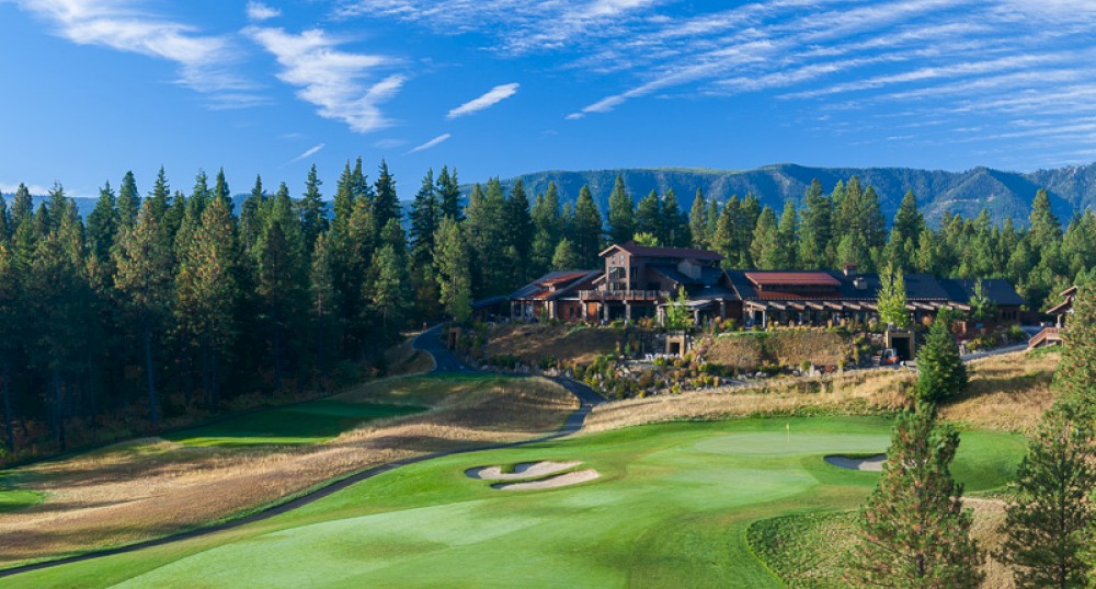 The image shows a scenic golf course with manicured greens, a clubhouse, and lush forested surroundings under a blue sky with scattered clouds.