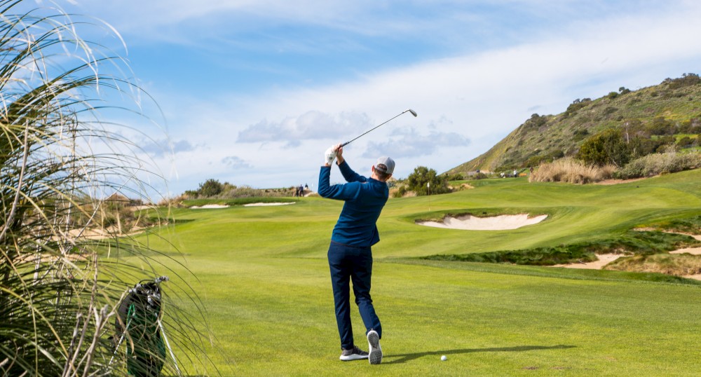 A person in blue attire is playing golf on a lush green golf course with sand bunkers, hills, and blue skies in the background.