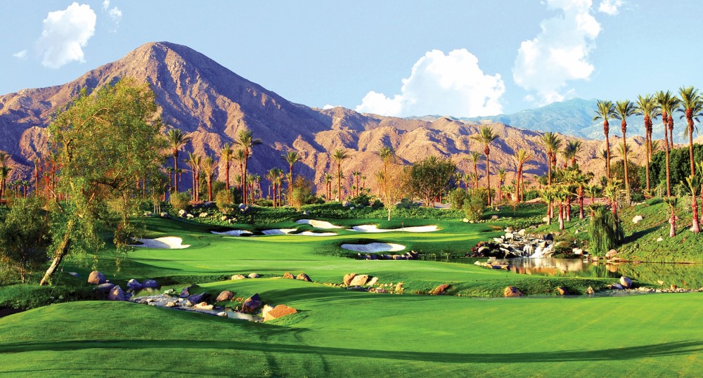 A picturesque golf course set against a backdrop of mountains, lush green fairways, palm trees, and clear skies with scattered clouds.