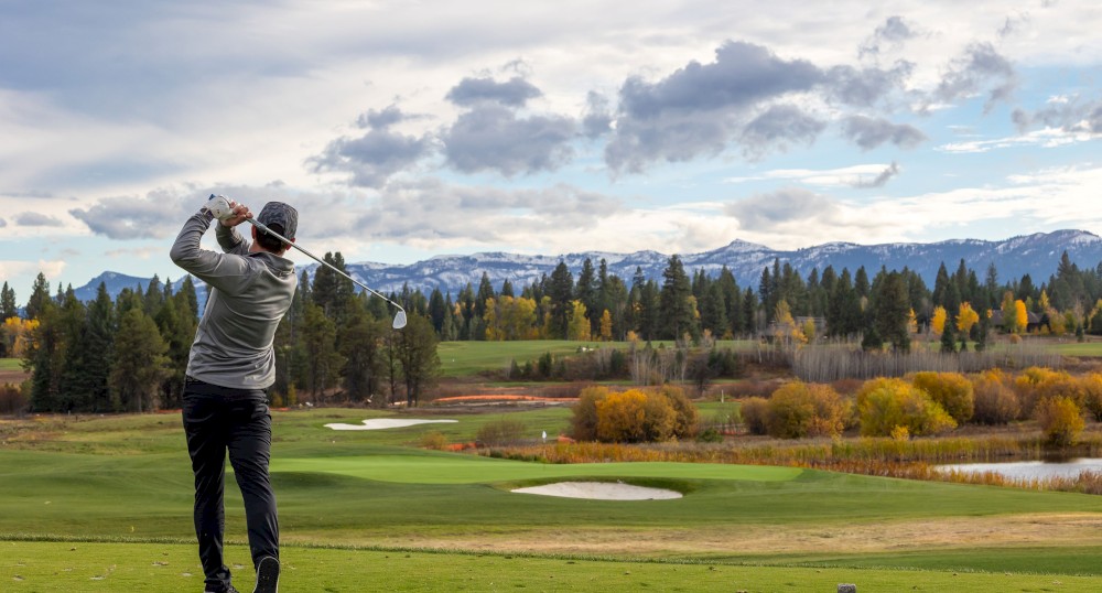 A golfer is mid-swing on a lush golf course with a scenic backdrop of mountains, trees in autumn colors, and a partly cloudy sky ending the sentence.