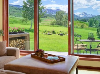 A cozy living room with a large window showing a scenic view of a green field, mountains, and a patio with outdoor furniture.