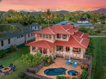 A large house with a red roof, surrounded by lush greenery, has a pool, lounge chairs, and an outdoor seating area, against a mountain backdrop.
