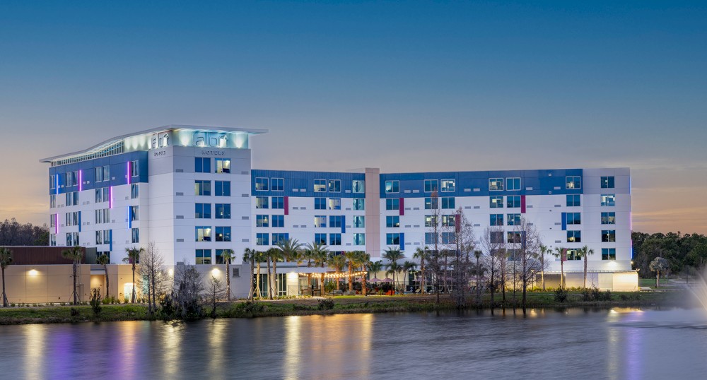 A modern multi-story building by a calm waterfront at dusk with a colorful facade, reflecting lights on the water