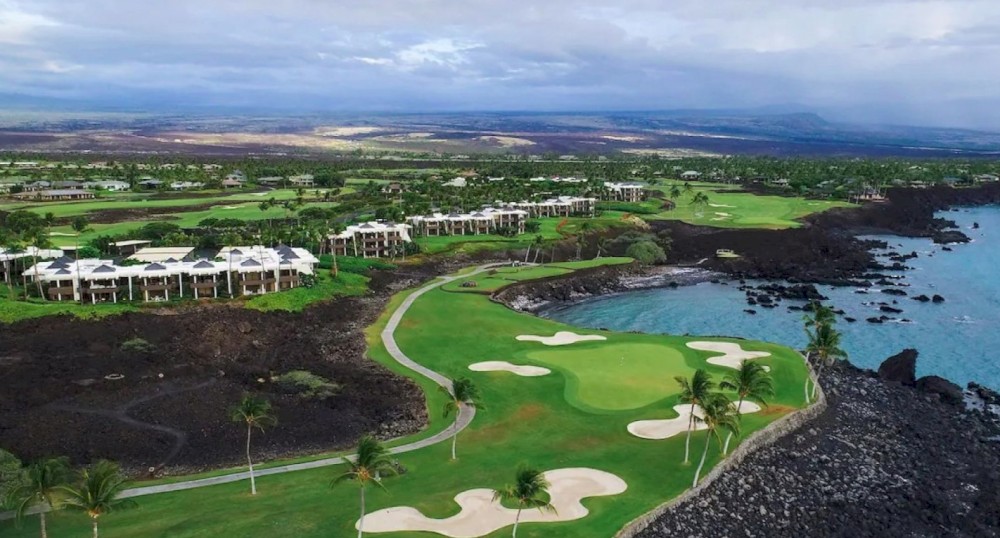 The image shows a coastal golf course surrounded by green landscapes, residential buildings, and the ocean with rocky shores, under a cloudy sky.