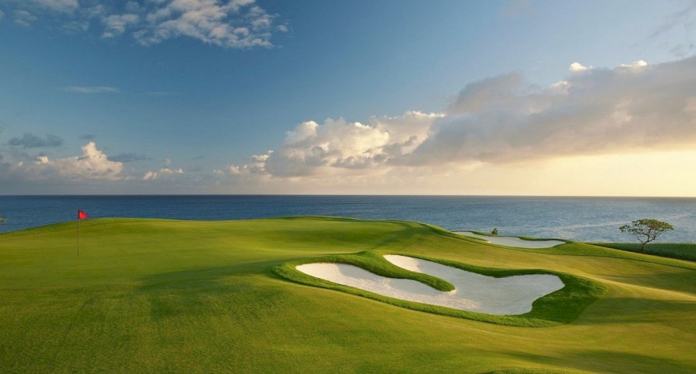 A scenic golf course near the ocean with a well-maintained green, sand bunkers, and a red flag under a partly cloudy sky at sunset.